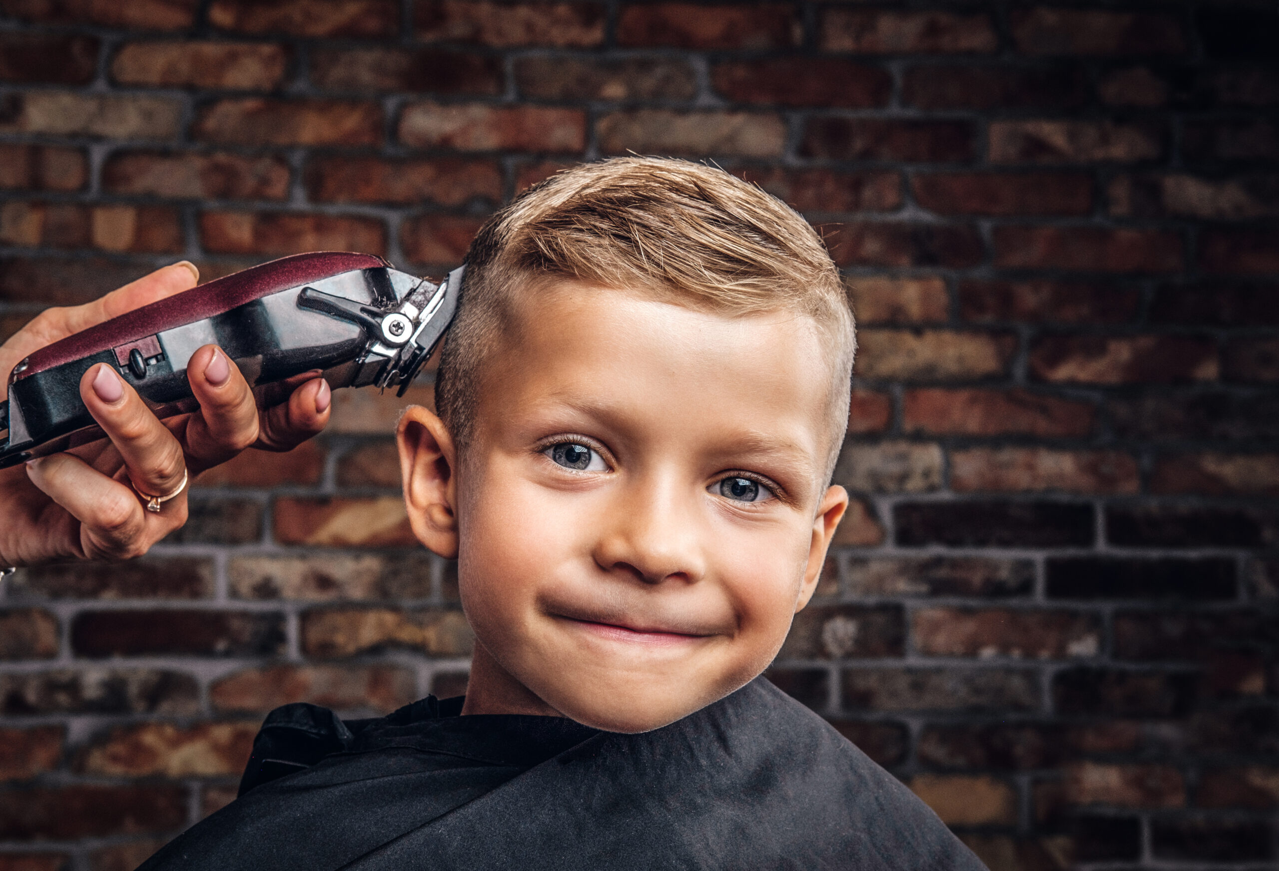 Close-up portrait of a cute smiling boy getting haircut against the brick wall.